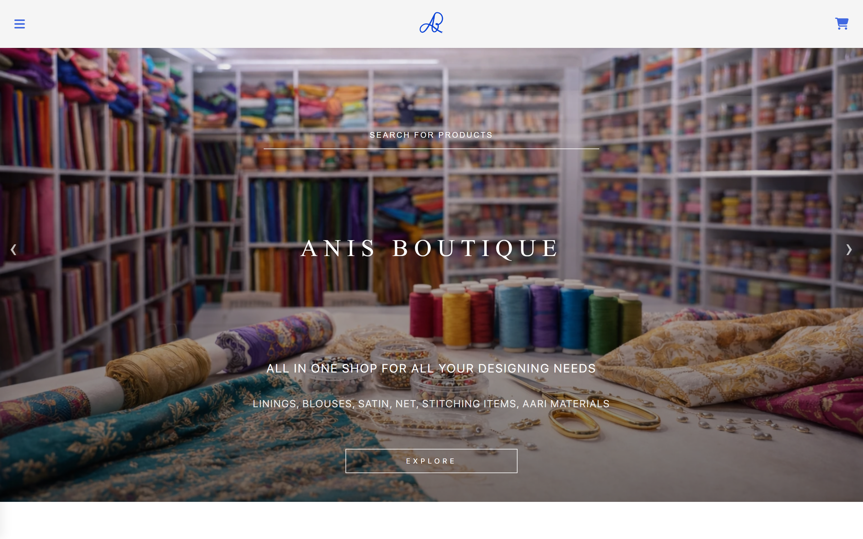 The Anis Boutique home page: a full-bleed photograph of the shop’s interior, its shelves stacked floor to ceiling with bolts of coloured fabric, and a worktop of thread spools, beading and gold scissors in the foreground. “Anis Boutique” is set across the middle above the lines “all in one shop for all your designing needs” and “linings, blouses, satin, net, stitching items, aari materials”, with a search field above and an Explore button below.